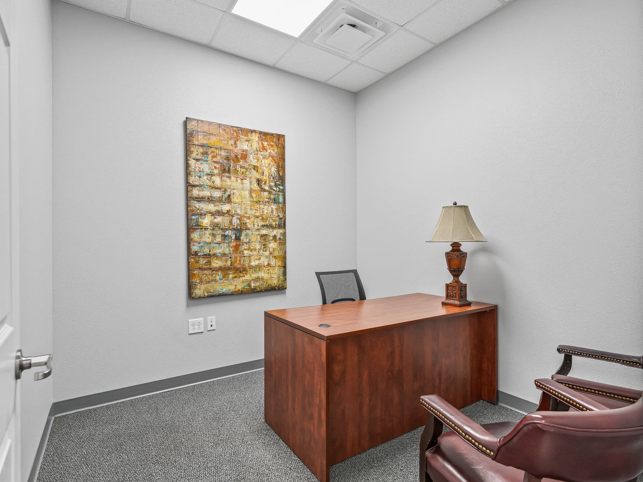 Small office with a wooden desk, a leather chair, and a mesh office chair. A decorative lamp sits on the desk. Abstract art hangs on a white wall.