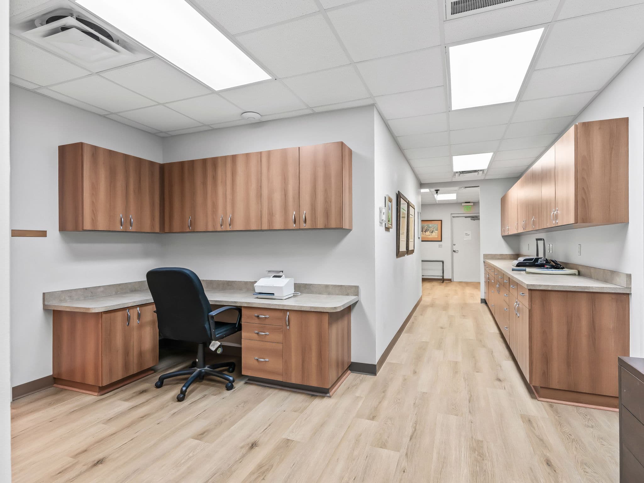 Spacious, modern office with wooden cabinets and light wood flooring. A black office chair sits at a desk with office equipment, creating a clean, professional ambiance.