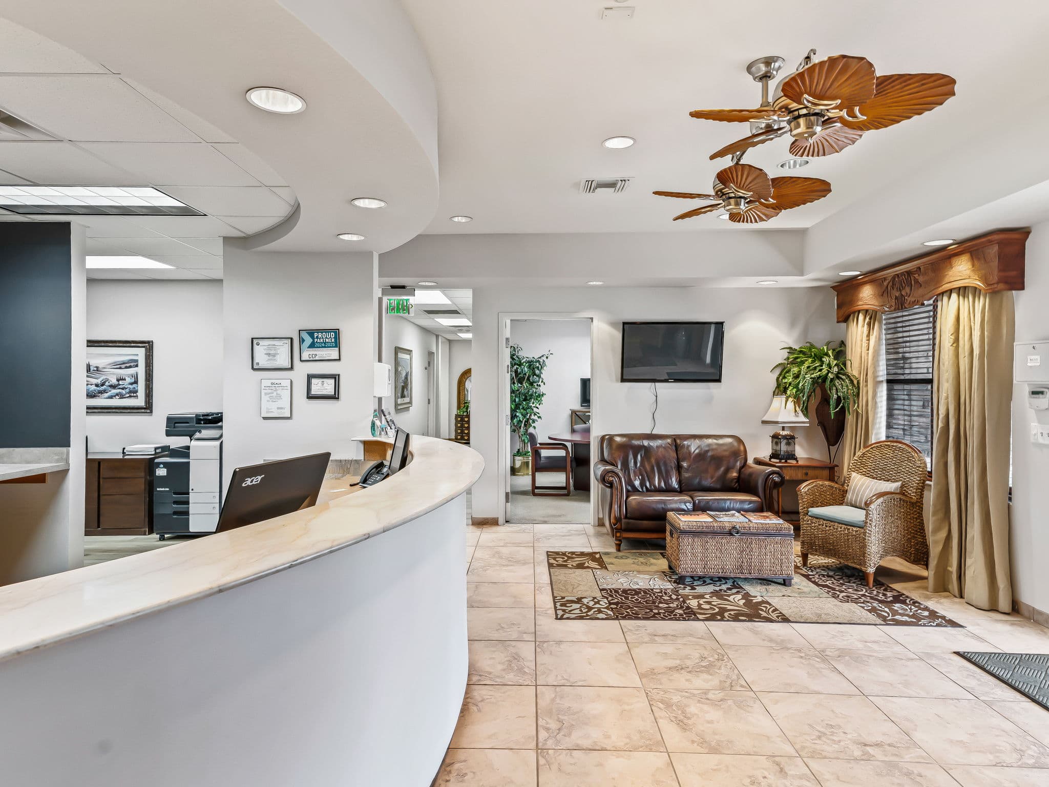 A bright office waiting area with a curved reception desk on the left. The room features a leather sofa, wicker chair, ceiling fans, and a potted plant.