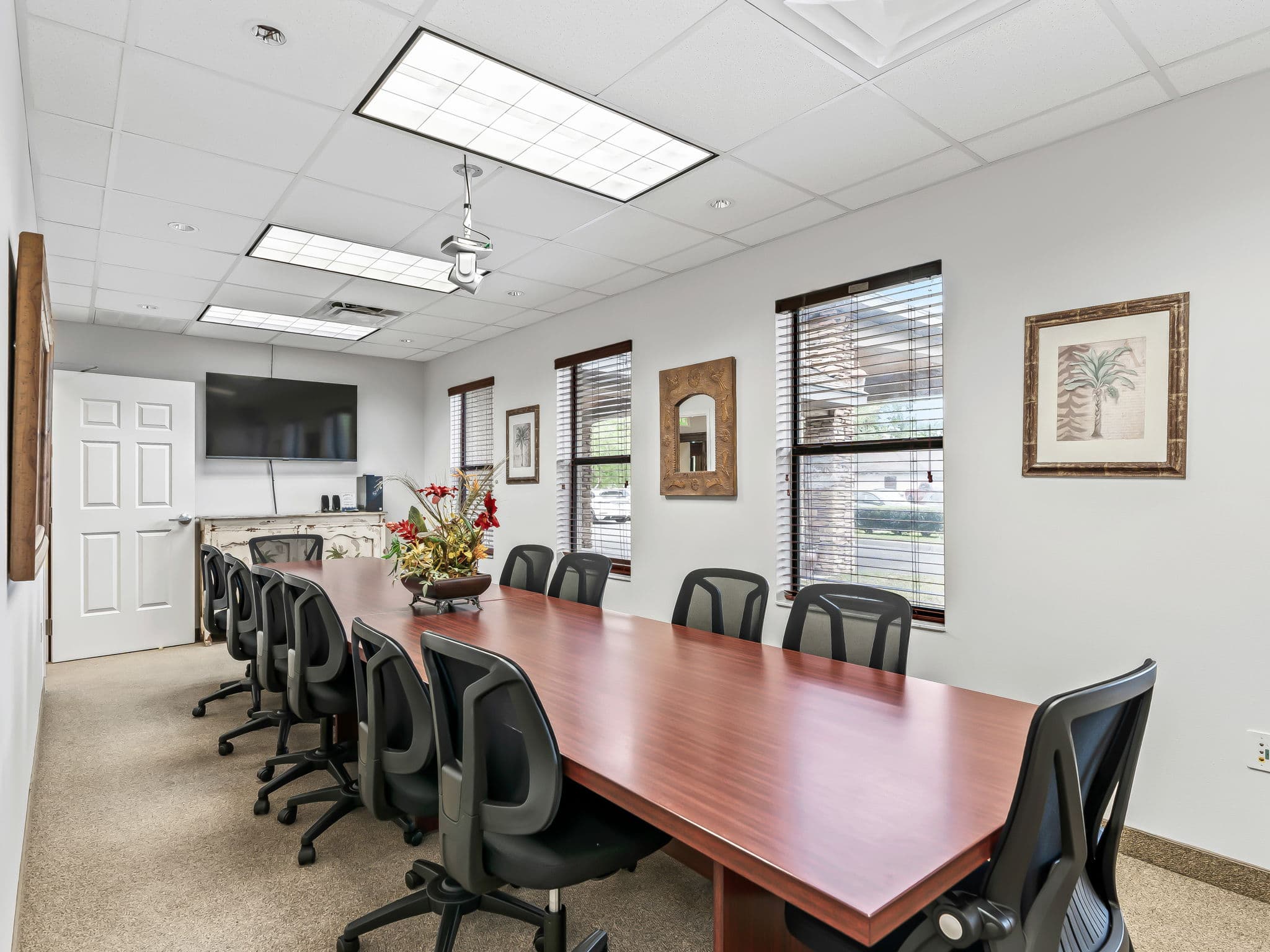 Modern conference room with a long wooden table, ten black swivel chairs, a wall-mounted TV, large windows with blinds, and decorative wall art.