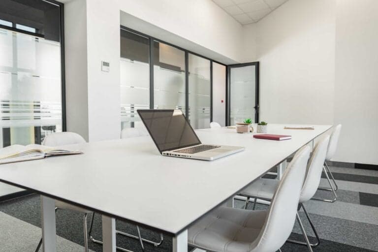 A sleek, bright meeting room at a premier office center featuring a long white conference table with a laptop, white tufted chairs, and frosted glass partitions under bright natural lighting.