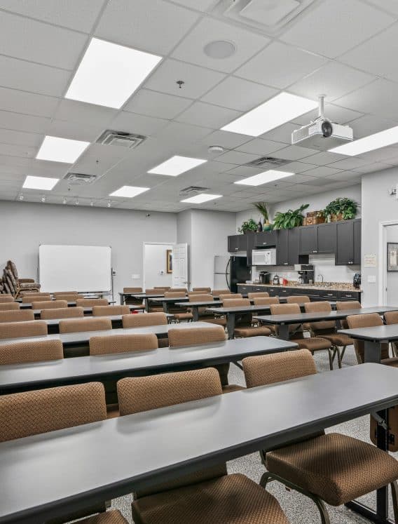 Copy of 18-_OpulentREMedia-18 A brightly lit classroom with rows of brown chairs and long tables facing a whiteboard and a projector. Cabinets with plants are at the back.