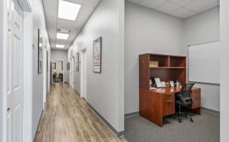 A modern office hallway with gray walls and wood flooring. On the right, there's a wooden desk with a black chair and a whiteboard, creating a clean, professional atmosphere.