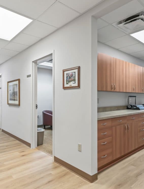 Spacious office hallway with wooden cabinets, framed artwork on white walls, and ceiling lights. Open doors reveal additional rooms, creating a professional atmosphere.
