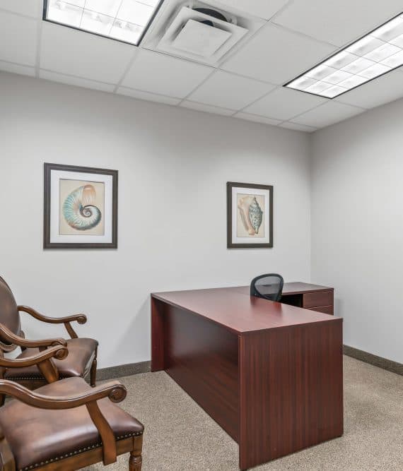 Plain office room with a wooden desk, a black chair, and two brown leather chairs. Walls are adorned with two nautical-themed artworks.