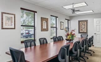 A long conference room with a wooden table, black chairs, and a floral centerpiece. Windows line one wall, and framed art decorates the light gray walls.