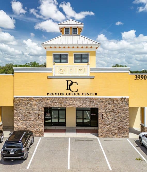 A yellow and tan two-story office building labeled Premier Office Center with stone accents, large windows, and four parked cars in front, under a partly cloudy sky. The address 3990 is visible on the right side.