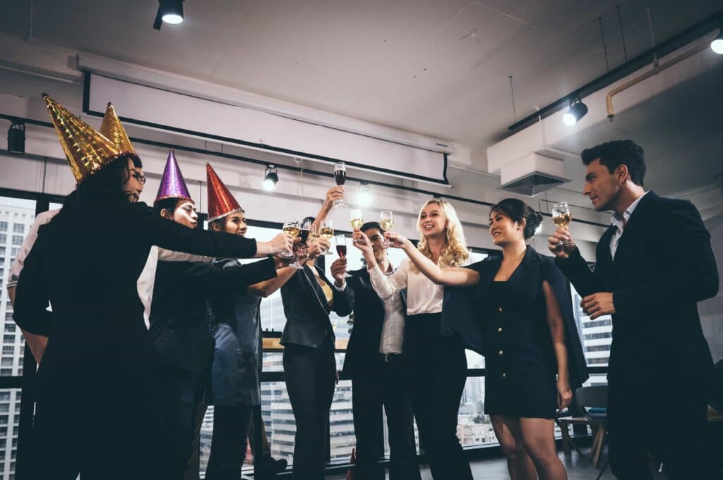 A diverse group of professional colleagues in business attire and festive party hats smiling and making a toast with champagne glasses during an evening office celebration with a city skyline view.