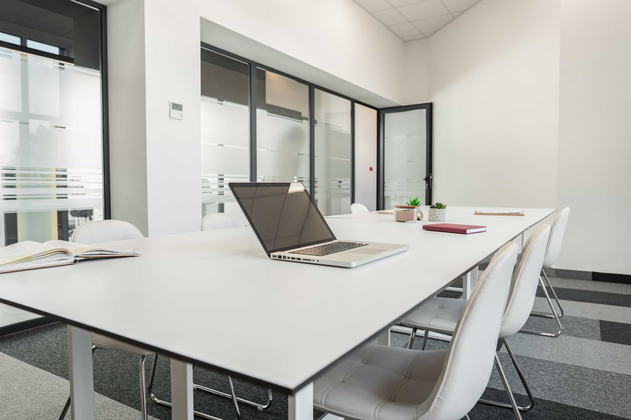 A sleek, bright meeting room at a premier office center featuring a long white conference table with a laptop, white tufted chairs, and frosted glass partitions under bright natural lighting.