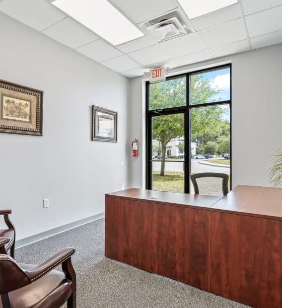 Simple office with a wooden desk, two brown chairs, wall art, and a large window showing greenery outside. A potted plant adds warmth to the space.
