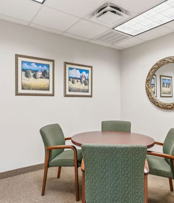 Copy of 14-_OpulentREMedia-34 Small meeting room with a round table and four green chairs. Two landscape paintings and a round, ornate mirror on the walls add a calm ambiance.