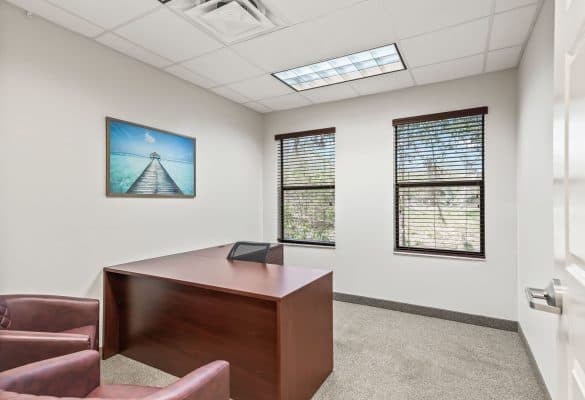 Large, bright office with a wooden desk, two maroon chairs, and a serene beach painting. Windows with blinds let in natural light, creating a calm atmosphere.