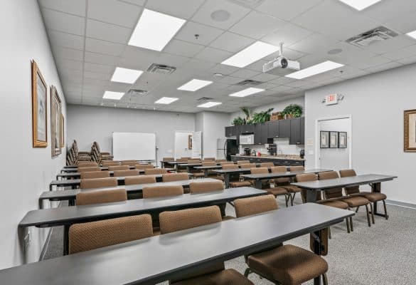 A brightly lit classroom with rows of brown chairs and long tables facing a whiteboard and a projector. Cabinets with plants are at the back.