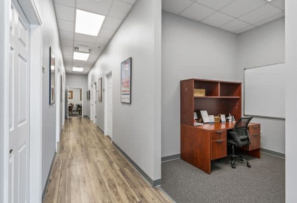 A modern office hallway with gray walls and wood flooring. On the right, there's a wooden desk with a black chair and a whiteboard, creating a clean, professional atmosphere.
