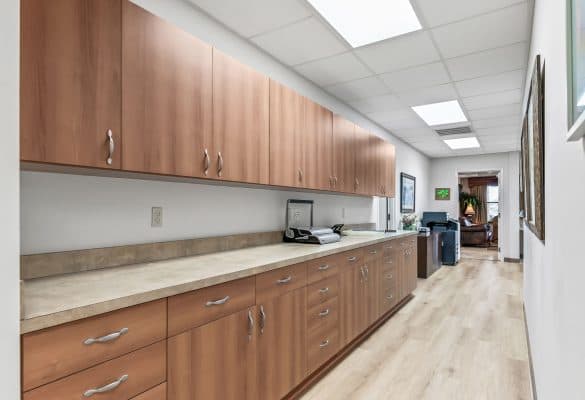 Copy of 23-_OpulentREMedia-9 Office hallway with wooden cabinets and beige countertop on the left, bright overhead lighting, light wood flooring, and a printer near the far end.