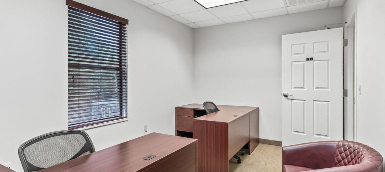 Copy of 35-_OpulentREMedia-21 Small, bright office with two wooden desks, mesh chairs, a window with blinds, and a wooden door. The room conveys a professional atmosphere.