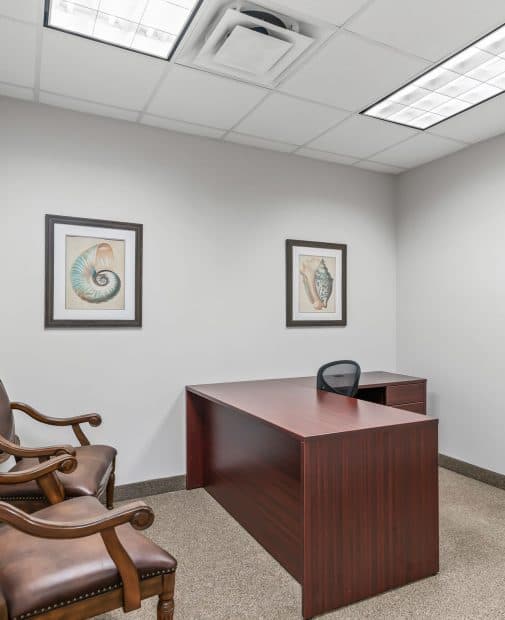 Small office with a wooden desk, black chair, and two leather armchairs. Walls feature framed seashell art, and fluorescent lighting brightens the space.