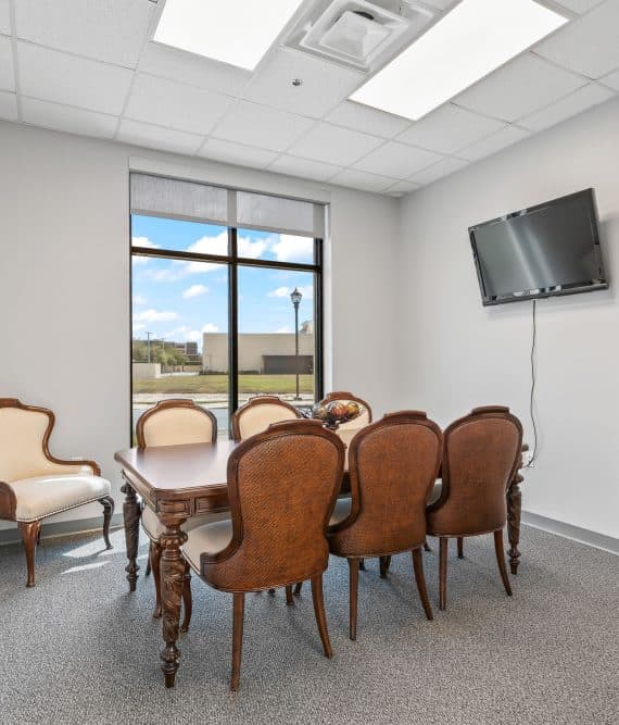 Copy of 6-_OpulentREMedia-4 A conference room with a wooden table and eight upholstered chairs, a wall-mounted TV, large window, artwork, and bright lighting.