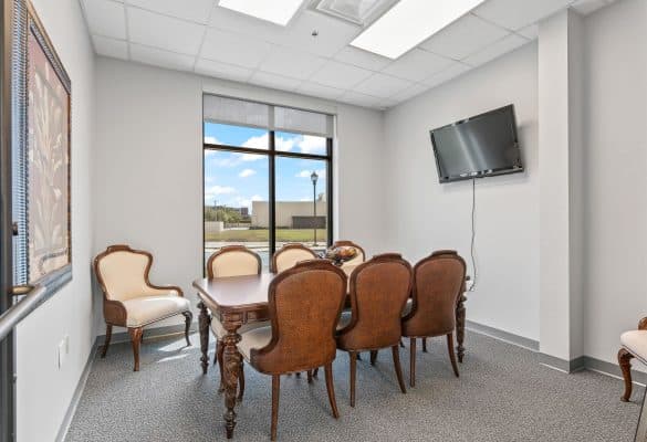 A conference room with a wooden table and eight upholstered chairs, a wall-mounted TV, large window, artwork, and bright lighting.