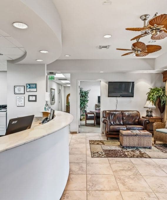 A bright office waiting area with a curved reception desk on the left. The room features a leather sofa, wicker chair, ceiling fans, and a potted plant.