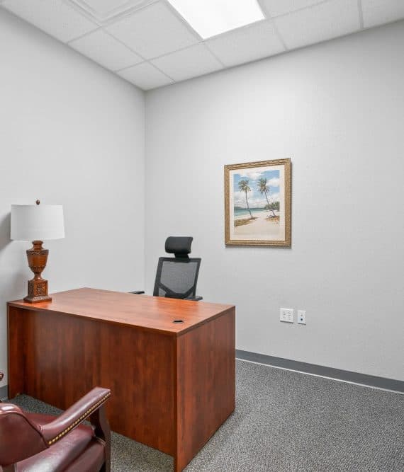 Small, minimalist office with a wooden desk, a black chair, and two brown leather chairs. A beach painting hangs on a white wall, under soft lighting.