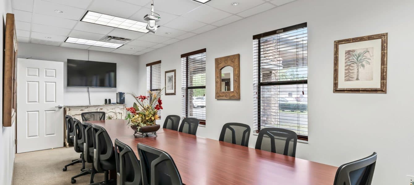 Copy of Copy of 10-_OpulentREMedia-23 Modern conference room with a long wooden table, ten black swivel chairs, a wall-mounted TV, large windows with blinds, and decorative wall art.