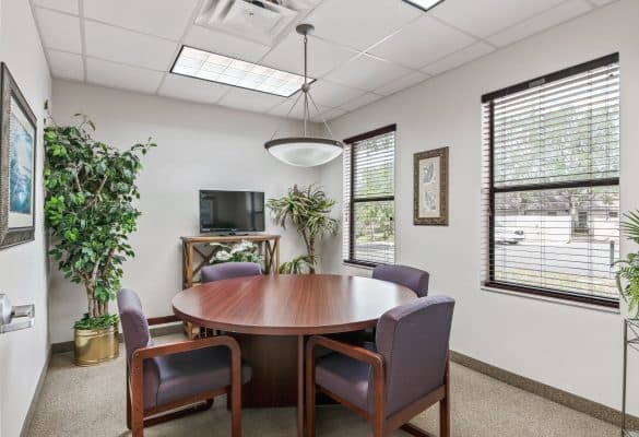 Copy of Copy of 9-_OpulentREMedia-36 A small office meeting room with a round wooden table and four purple chairs. Two large windows with blinds offer natural light. Green plants add a calming touch.