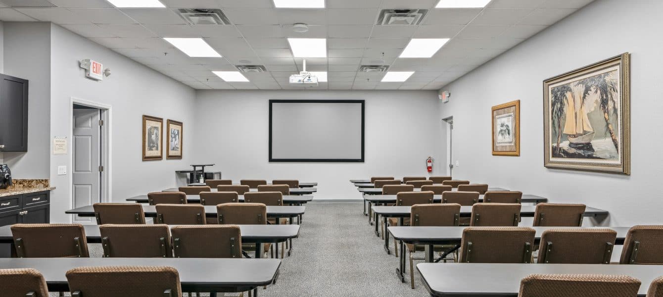 A well-lit conference room with rows of empty chairs and tables facing a blank projection screen. Framed artwork adorns the light gray walls.